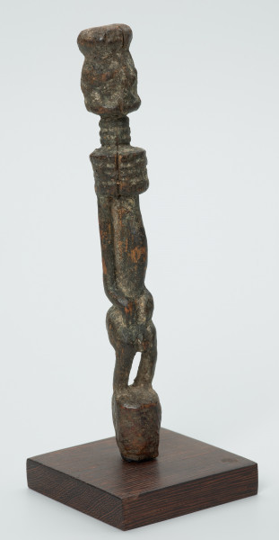 rzeźba - Ujęcie przodu skosem w prawą stronę. Drewniana rzeźba postaci mężczyzny ze zgietymi kolanami. Schematyczne zarysowanie dużej głowy, długiej szyi, na której widoczny jest szeroki naszyjnik. Wąski, długi tułów, długie ręce oparte na kolanach, krótkie nogi i szerokie stopy.