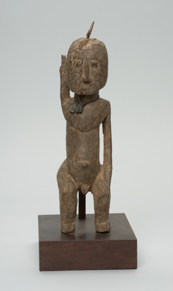rzeźba - Ujęcie z przodu. Figura przedstawiająca mężczyznę, o czym świadczy lekko zaznaczony penis. Prawa ręka krótsza od lewej, uniesiona do góry. Ramiona proste, pośladki i uda zaokrąglone. Na głowie wbity kawałek żelaza. Związana z kultem przodków.