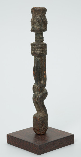 rzeźba - Ujęcie przodu skosem w lewą stronę. Drewniana rzeźba postaci mężczyzny ze zgietymi kolanami. Schematyczne zarysowanie dużej głowy, długiej szyi, na której widoczny jest szeroki naszyjnik. Wąski, długi tułów, długie ręce oparte na kolanach, krótkie nogi i szerokie stopy.