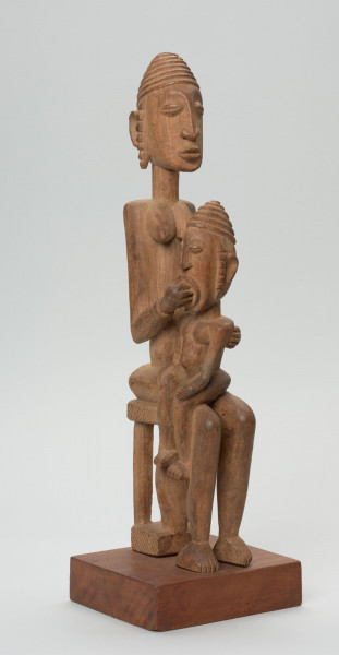 rzeźba - Ujęcie przodu i boku; Figurka przedstawiająca postać kobiety siedzącej na stołu i trzymającej na kolanach dziecko. Kobieta ma owalną, wydłużoną twarz oraz długie, zwisające uszy. Rysy twarzy proporcjonalne, z cienkim kształtnym nosem, brwiami oraz wypukłymi oczami i ustami.