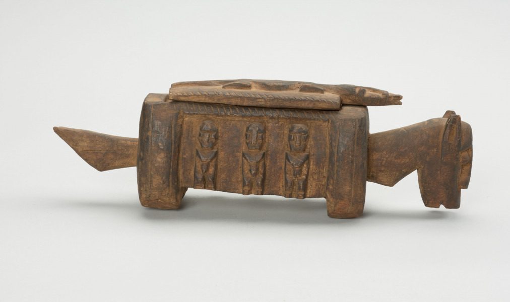 rzeźba - Ujęcie z prawego boku; Arka Nommo zwana inaczej aduno koro lub arką świata, jest wyobrażeniem mitycznej łodzi. Drewniana rzeźa przypominająca konia. Bok zdobiony trzema postaciami w formie płaskorzeźby, stojące w rzędzie. Głowa obiektu zwrócona w stronę ziemi. Na grzbiecie druga część obiektu w formie przykrywki kształtem przypominająca krokodyla na drwnianej prostokatnej podstawie.