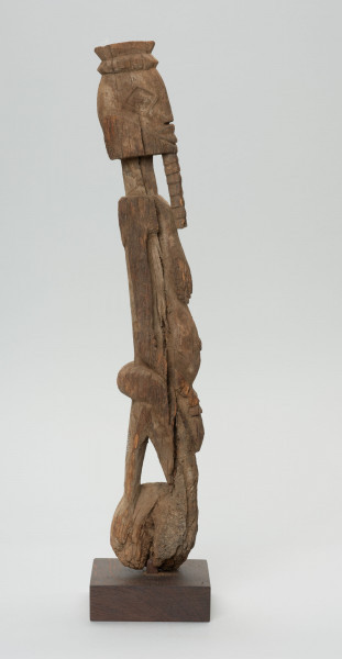 rzeźba - Ujęcie z lewego boku; Figura - postać kobiety  - przodka, obdarzona została bowiem ozdobną brodą, czyli pewną cechą męską. Głowa i korpus oddzielone od szyi prostymi cięciami. Oczy duże wypukłe, piersi długie, płaskie, nogi zatarte w wyniku używania. Związana z kultem przodków.