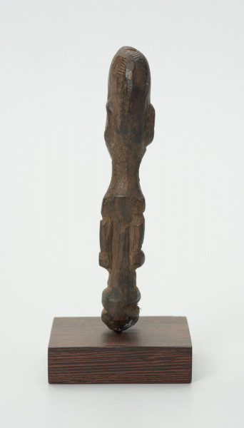 Drewniana figurka z dużą głową i wąskimi oczami - Ujęcie z tyłu; mała drewniana figurka antropomorficzna. Głowa duża zaokrąglona. Rysy, ręce wydobyte z bryły drewna płytkimi cięciami. Brak nóg.