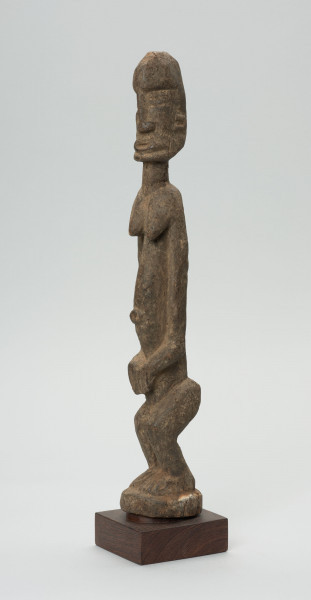 rzeźba - Ujęcie ze skosu z prawej; Figura - postać kobiety w pozycji stojącej. Głowa oddzielona od szyi prostą linią. Okrągłe czoło, nos długi, prosty, strzałkowaty, usta i oczy szerokie. Ręce wydobyte płytkimi nacięciami z bryły korpusu. Dłonie spoczywają na łonie. Nogi ugięte w kolanach. Zaznaczone stopy.