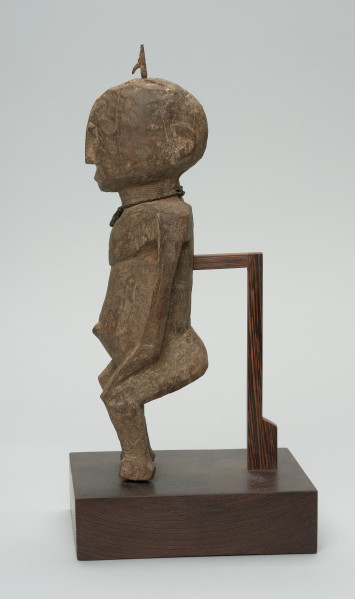 rzeźba - Ujęcie lewego boku. Figura przedstawiająca mężczyznę, o czym świadczy lekko zaznaczony penis. Prawa ręka krótsza od lewej, uniesiona do góry. Ramiona proste, pośladki i uda zaokrąglone. Na głowie wbity kawałek żelaza. Związana z kultem przodków.