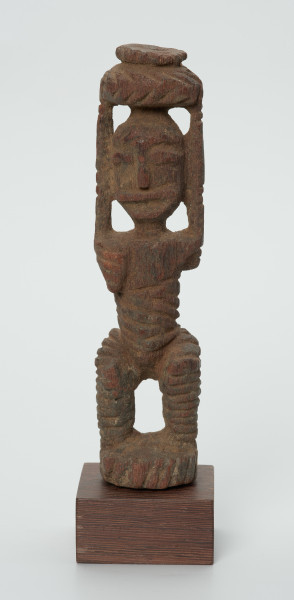 drewniana figura kobiety - Ujęcie z przodu. Drewniana, rzeźbiona figura kobiety niosącej na głowie naczynie. Postać schematyczna, z uniesionymi ramionami. Głowa jajowata, rysy twarzy uproszczone. Stopy złączone w okrągły cokolik.