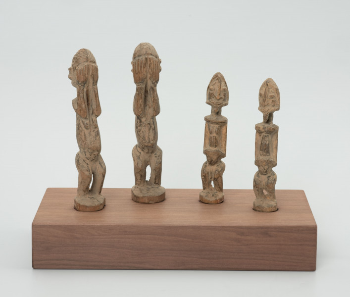 rzeźba - Ujęcie z przodu; Zestaw czterech jasnych, drewnianych figurek przedstawiających postacie ludzkie w pozycji stojącej – dwie kobiety i dwóch mężczyzn. Większa para ma zakryte dłońmi oczy. Rysy twarzy – w postaci oczu, strzałkowego nosa, ust i brody – zaznaczono na mniejszych rzeźbach, których ręce przylegają do tułowia, a dłonie oparto o lekko wypukły i wystający brzuch. Wszystkie rzeźby mają krótkie, ugięte w kolanach nogi. Nie zaznaczono im stóp, w ich miejscu wyrzeźbiono okrągłe podstawki. Wszystkie figurki zostały umieszczone przez konserwatora na prostokątnej, drewnianej, ekspozycyjnej podstawie.