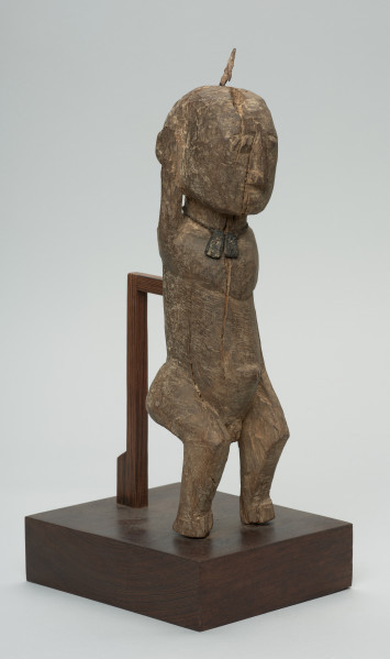 rzeźba - Ujęcie z przodu z prawej strony. Figura przedstawiająca mężczyznę, o czym świadczy lekko zaznaczony penis. Prawa ręka krótsza od lewej, uniesiona do góry. Ramiona proste, pośladki i uda zaokrąglone. Na głowie wbity kawałek żelaza. Związana z kultem przodków.