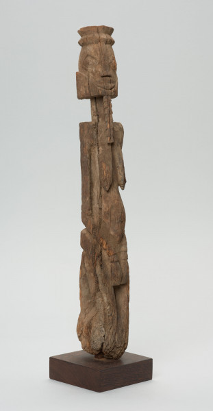 rzeźba - Ujęcie ze skosu z lewej; Figura - postać kobiety  - przodka, obdarzona została bowiem ozdobną brodą, czyli pewną cechą męską. Głowa i korpus oddzielone od szyi prostymi cięciami. Oczy duże wypukłe, piersi długie, płaskie, nogi zatarte w wyniku używania. Związana z kultem przodków.