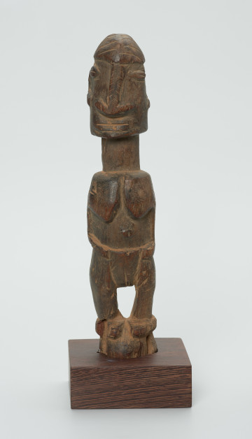 Drewniana figurka kobiety z szeroko rozstawionymi oczami - Ujęcie z przodu. Drewniana figurka kobiety w pozycji stojącej. Oczy szeroko rozstawione, zaznaczony wąski nos, rozszerzony na dole, szerokie usta. Zaznaczone płaskie piersi oraz narządy rozrodcze. Stopy złączone ze sobą w formie niewielkiego cokoliku.