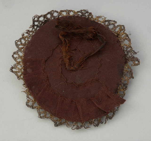 rozeta do obuwia księcia Franciszka I (1577-1620) - Ujęcie spodu pierwszej rozety. Rozeta wykonana z wykończonej złotą koronką jedwabnej wstążki, ułożonej spiralnie i przyszytej do okrągłej podkładki. Spięta pośrodku trójlistnym klejnotem zdobionym barwną emalią. Na odwrocie rozety zachowane fragmenty wstążek.
