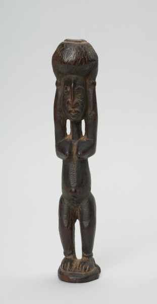 rzeźba - Ujęcie z przodu. Figura - postać kobiety w pozycji stojącej z rękoma wyciągniętymi ku górze, którymi podtrzymuje stojące na głowie naczynie. Na brzuchu delikatne nacięcia - skaryfikacja. Zaznaczone stopy. Przy dłoniach i stopach - okrągłe zgrubienia - wizerunki bransolet.