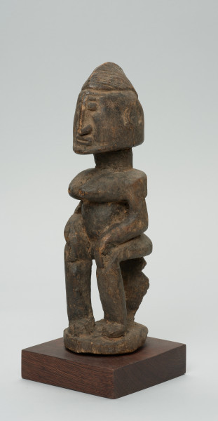 rzeźba - Ujęcie przodu skosem w lewą stronę. Figura przedstawiająca postać kobiety siedzącej na stołku. Postać masywna, głowa nieproporcjonalnie duża, ramiona proste, ręce przylegające do tułowia, dłonie spoczywają na kolanach. Nos długi, prosty, w kształcie strzałki, oczy i usta zaznaczone symbolicznie.