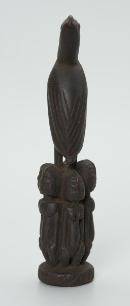 Drewniana figurka ptaka na cokole z postaci ludzkich - Ujęcie z tyłu. Drewniana figurka ptaka ze skrzydłami ułożonymi wzdłuż tułowia, stojącego na głowach zwróconych do siebie plecami pięciu postaci ludzkich. Postaci, tworzące cokół, przedstawione statycznie, w formie zgeometryzowanej.