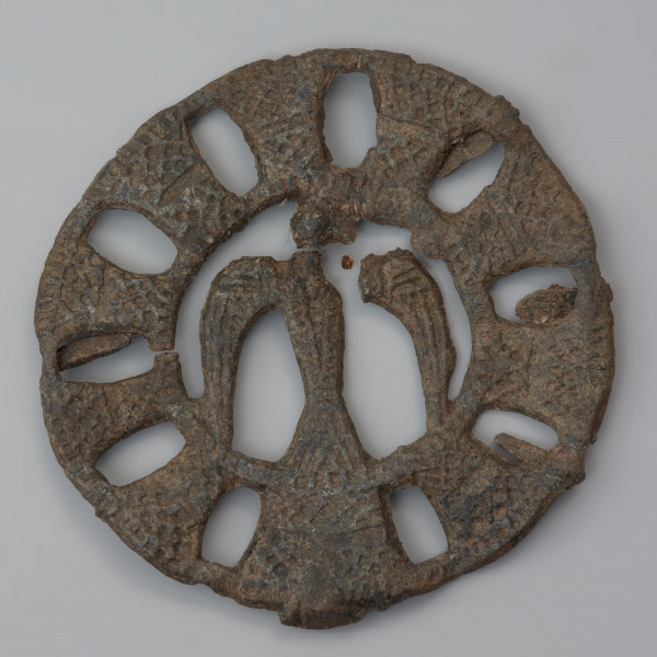 plakietka ażurowa z ptakiem - Ujęcie przodu z góry. Metalowa, okrągła plakietka ażurowa z wyobrażeniem ptaka w części centralnej, otoczonego koncentrycznymi kręgami z owalnymi otworami nadającymi całości pewnej lekkości. Na odwrocie plastyczne zeberka ułożone promieniście, wzmacniające konstrukcję.