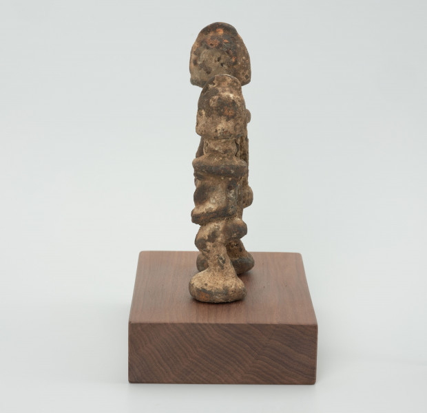 rzeźba - Ujęcie z lewego boku; Zestaw dwóch małych, drewnianych figurek postaci ludzkich w pozycji stojącej, pokrytych warstwą białej i miedzianej substancji. Figurki mają fryzury lub nakrycia głowy postawione na czubku głowy. Widoczne niewyraźne, okrągłe oczy i linia nosa. Ręce rzeźb  przylegają do tułowia, ich dłonie oparto o lekko wypukły i wystający brzuch, nogi krótkie i ugięte w kolanach, stopy połączone, stoją na okrągłych podstawkach. Obie figurki zostały umieszczone przez konserwatora na wspólnej prostokątnej, drewnianej, ekspozycyjnej podstawie.