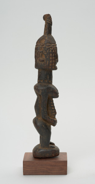 rzeźba - Ujęcie lewej strony rzeźby. Drewniana figura postaci kobiety. Podłużna głowa ze scheycznie zaznaczonymi rysami i ozdobną wysoką fryzurą. Długa masywna szyja, szczupły tułów. Wyraźnie zaznaczone piersi, długi ręce splecione z przodu. Postać ma krótkie nogi ugięte w kolanach.