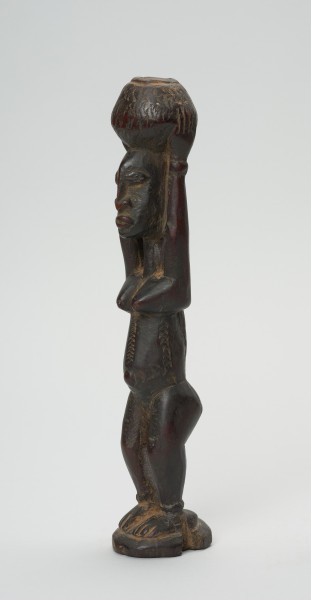 rzeźba - Ujęcie z przodu z lewej strony. Figura - postać kobiety w pozycji stojącej z rękoma wyciągniętymi ku górze, którymi podtrzymuje stojące na głowie naczynie. Na brzuchu delikatne nacięcia - skaryfikacja. Zaznaczone stopy. Przy dłoniach i stopach - okrągłe zgrubienia - wizerunki bransolet.