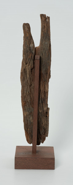 Drewniana figurka na rozwidlonej u góry desce - Ujęcie z tyłu; na płaskiej, rozwidlonej na szczycie desce wyrzeźbiono okrągłą głowę z delikatnie zaznaczonymi oczami, nosem z małymi ustami. Figura posiada liczne spękania.