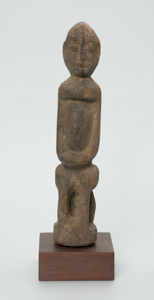 Drewniana figurka postaci siedzącej na stołku - Ujęcie z przodu; postać siedząca na stołku. Części ciała i twarz schematyczne, zaznaczone płytkimi nacięciami. Figura statyczna. Przez całą twarz pęknięcie sięgające aż do klatki piersiowej.