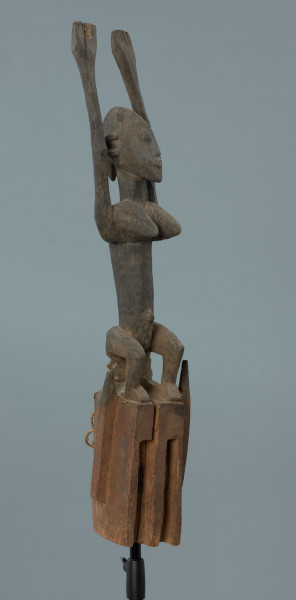 drewniana maska - Ujęcie z przodu, z lewej strony; Drewniana, rzeźbiona maska. Nad częścią twarzową, uproszczoną, z wywierconymi dziurami w miejscu oczu, figura postaci ludzkiej z rękoma wzniesionymi do góry. Głowa jajowata, spiczasto zakończona. Zaznaczone uszy. Na wąskim tułowiu wyeksponowane piersi.