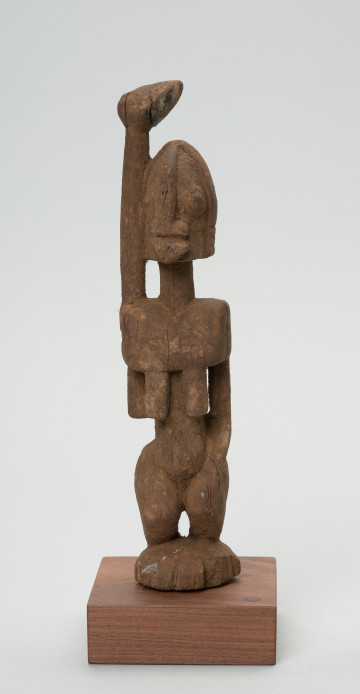 rzeźba - Ujęcie z przodu. Figura - postać kobiety z uniesioną ku górze prawą ręką. Całość uproszczona, zwarta. Piersi długie, płaskie - wskazują na to, że jest to wizerunek kobiety starej. Związana z kultem przodków, przeznaczona na ołtarz przodków.