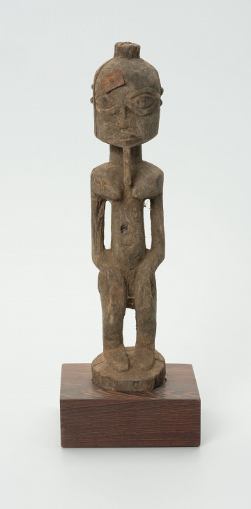 Figurka postaci z długą cienką brodą - Ujęcie z przodu; Figurka postaci z długą cienką brodą. Duża, zaokrąglona głowa, zakończona długą, cienką bródką. Oczy duże ze źrenicami. Ręce wyodrębnione z korpusu. Całość statyczna. Na głowie umieszczony kawałek metalu