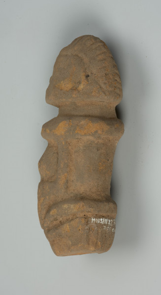 Gliniana figurka kobiety z wypukłym brzuchem - Ujęcie tyłu lewej strony; obiekt leżący na płasko; gliniana figurka kobiety siedzącej na podkulonych nogach. Głowa duża, oddzielona od szyi, prostą linią, barki proste. Brzuch wypukły, uformowany stożkowato.  Ręce i nogi lekko wypukłe.