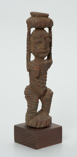 drewniana figura kobiety - Ujęcie z przodu skosem w prawą stronę. Drewniana, rzeźbiona figura kobiety niosącej na głowie naczynie. Postać schematyczna, z uniesionymi ramionami. Głowa jajowata, rysy twarzy uproszczone. Stopy złączone w okrągły cokolik.