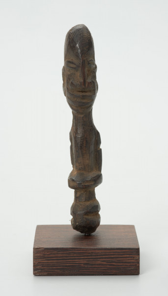 Drewniana figurka z dużą głową i wąskimi oczami - Ujęcie z przodu; mała drewniana figurka antropomorficzna. Głowa duża zaokrąglona. Wąskie oczy. Rysy, ręce wydobyte z bryły drewna płytkimi cięciami. Brak nóg.