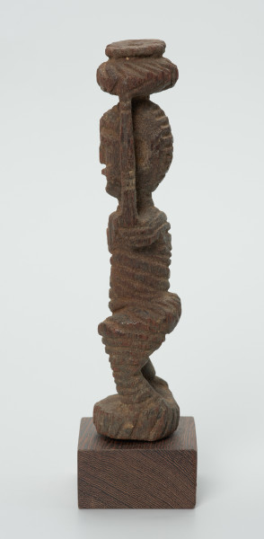 drewniana figura kobiety - Ujęcie prawego boku. Drewniana, rzeźbiona figura kobiety niosącej na głowie naczynie. Postać schematyczna, z uniesionymi ramionami. Głowa jajowata, rysy twarzy uproszczone. Stopy złączone w okrągły cokolik.