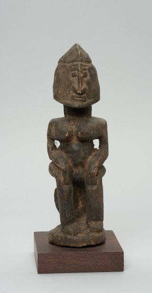 rzeźba - Ujęcie z przodu. Figura przedstawiająca postać kobiety siedzącej na stołku. Postać masywna, głowa nieproporcjonalnie duża, ramiona proste, ręce przylegające do tułowia, dłonie spoczywają na kolanach. Nos długi, prosty, w kształcie strzałki, oczy i usta zaznaczone symbolicznie.