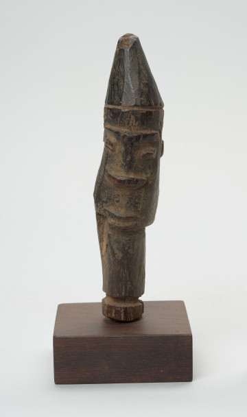 rzeźba - Ujęcie z przodu. Figura - głowa zakończona uchwytem. Głowa z bardzo schematycznie przedstawionymi oczami, nosem i ustami. Na głowie spiczasta czapka. Szyja przechodzi w uchwyt. Związana z kultem przodków, przeznaczona na ołtarz przodków.