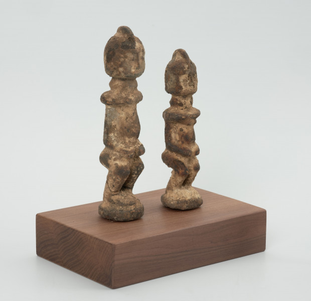 rzeźba - Ujęcie ze skosu z lewej; Zestaw dwóch małych, drewnianych figurek postaci ludzkich w pozycji stojącej, pokrytych warstwą białej i miedzianej substancji. Figurki mają fryzury lub nakrycia głowy postawione na czubku głowy. Widoczne niewyraźne, okrągłe oczy i linia nosa. Ręce rzeźb  przylegają do tułowia, ich dłonie oparto o lekko wypukły i wystający brzuch, nogi krótkie i ugięte w kolanach, stopy połączone, stoją na okrągłych podstawkach. Obie figurki zostały umieszczone przez konserwatora na wspólnej prostokątnej, drewnianej, ekspozycyjnej podstawie.