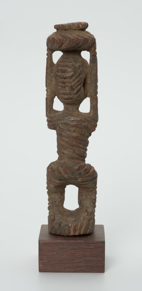 drewniana figura kobiety - Ujęcie z tyłu. Drewniana, rzeźbiona figura kobiety niosącej na głowie naczynie. Postać schematyczna, z uniesionymi ramionami. Głowa jajowata, rysy twarzy uproszczone. Stopy złączone w okrągły cokolik.