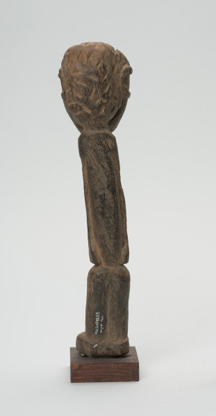 rzeźba - Ujęcie tyłu. Figurka przedstawiająca postać ludzką. Głowa bardzo duża, reszta sylwetki schematyczna, bardzo smukła, lekko wygięta w prawą stronę. Włosy zaznaczone ciosanymi rytami. Nogi wąskie, złączone.  W dolnej części rzeźby widnieje numer inwentarzowy.