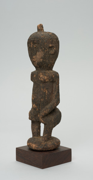 rzeźba - Ujęcie przodu skosem w lewą stronę. Figurka - postać kobiety w pozycji stojącej. Głowa duża wychodząca bezpośrednio z tułowia. Ręce przylegające do tułowia. Na głowie mała wypustka.