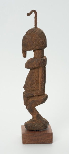 rzeźba - Ujęcie z prawej strony. Drewniana rzeźba postaci męskiej. Wąska podłużna twarz, na głowie haczykowata ozdoba. Tułów długi, nogi zgięte w kolanach, ręce oparte na kolanach.