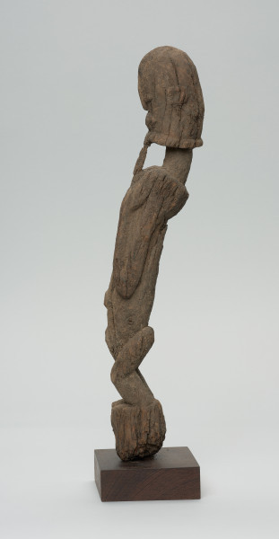 rzeźba - Ujęcie lewego boku. Figura antropomorficzna. Rysy zatarte, widoczna broda, wypukłe, okrągłe oczy. Korpus wydłużony, ręce przylegające do tułowia. Pośladki zaokrąglone. Postać stoi na małym, zaokrąglonym cokoliku.