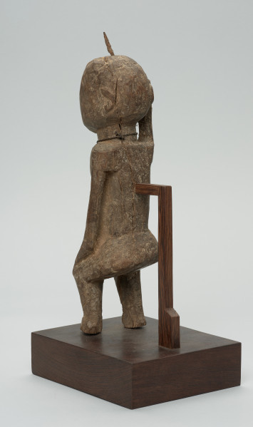 rzeźba - Ujęcie z tyłu z lewej strony. Figura przedstawiająca mężczyznę, o czym świadczy lekko zaznaczony penis. Prawa ręka krótsza od lewej, uniesiona do góry. Ramiona proste, pośladki i uda zaokrąglone. Na głowie wbity kawałek żelaza. Związana z kultem przodków.