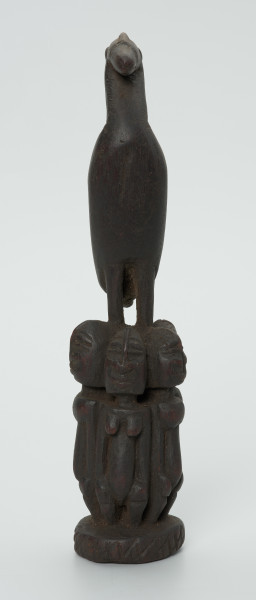 Drewniana figurka ptaka na cokole z postaci ludzkich - Ujęcie z przodu. Drewniana figurka ptaka ze skrzydłami ułożonymi wzdłuż tułowia, stojącego na głowach zwróconych do siebie plecami pięciu postaci ludzkich. Postaci, tworzące cokół, przedstawione statycznie, w formie zgeometryzowanej.