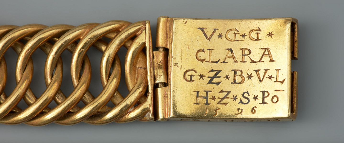 Bransoleta z sarkofagu księcia Franciszka I (1577 - 1620) - Ujęcie prawego zapięcia. Bransoleta wykonana ze skręconych, przenikających się ogniw, w tzw. splocie pancerzowym. Na prostokątnych zapinkach dziewięciopolowy herb Księstwa Pomorskiego trzymany przez dzikich mężów, pierwotnie emaliowany. Na spodzie klamry ryta inskrypcja, pierwotnie emaliowana: V.G.G. / CLARA / G.Z.B.V.L. / H.Z.S.Pō /15.96. Na bocznych ściankach wykonana w tej samej technice inskrypcja: V.G.G. FRANTZ / H.Z.S.Pom.