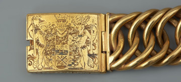bransoleta z sarkofagu księcia Franciszka I (1577 - 1620) - Ujęcie zapięcia z lewej. Bransoleta wykonana ze skręconych, przenikających się ogniw, w tzw. splocie pancerzowym. Na prostokątnych zapinkach dziewięciopolowy herb Księstwa Pomorskiego trzymany przez dzikich mężów, pierwotnie emaliowany. Na spodzie klamry ryta inskrypcja, pierwotnie emaliowana: V.G.G. / CLARA / G.Z.B.V.L. / H.Z.S.Pō /15.96. Na bocznych ściankach wykonana w tej samej technice inskrypcja: V.G.G. FRANTZ / H.Z.S.Pom.
