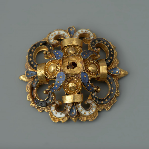 rozeta - Ujęcie z przodu. Ozdoba w kształcie ażurowej rozety. Wokół centralnie umieszczonego złotego guza rozmieszczone są cztery mniejsze guzy otoczone filigranem, płatki zdobione ciemnoniebieską emalią, a także woluty wypełnione czarną oraz białą emalią, spięte złotymi taśmami. Elementy emaliowane są inkrustowane regularnie rozmieszczonymi złotymi kropkami. Na odwrocie uszko służące do przyszywania rozet do strojów.
