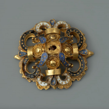 rozeta - Ujęcie z przodu. Ozdoba w kształcie ażurowej rozety. Wokół centralnie umieszczonego złotego guza rozmieszczone są cztery mniejsze guzy otoczone filigranem, płatki zdobione ciemnoniebieską emalią, a także woluty wypełnione czarną oraz białą emalią, spięte złotymi taśmami. Elementy emaliowane są inkrustowane regularnie rozmieszczonymi złotymi kropkami. Na odwrocie uszko służące do przyszywania rozet do strojów.