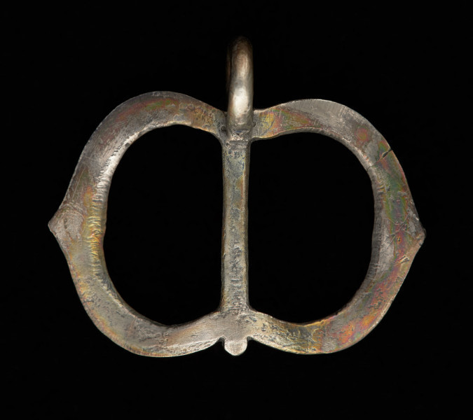 klamra od pendentu - Ujęcie z tyłu. Rama klamry zbudowana z dwóch przedzielonych gładkim trzpieniem części, które w zewnętrznym obrysie mają kształt łuku w ośli grzbiet, a od wewnątrz łuku koszowego. Przednie i boczne ścianki ramy zdobione są rytym, niellowanym ornamentem. U dołu gładkie, okrągłe uszko, mocowane poprzecznie.