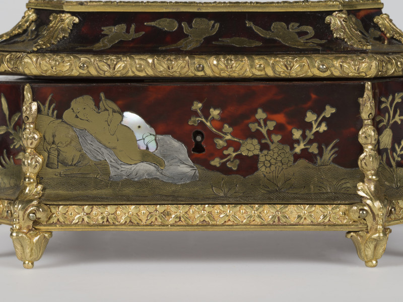 zbliżenie na dekorację z przodu - Amor leżący, widok z boku, motyw roślinny po prawej,  w środku dziurka na klucz, krawędzie szkatułki pokryte dekoracjami z brązu w postaci palmet i ornamentu kwiatowego, nóżki szkatułki w postaci rozety liści.