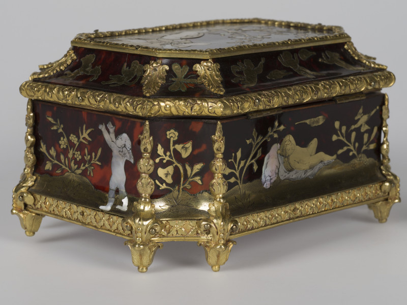zbliżenie na dekorację prawego tylnego narożnika, widok z boku, motyw roślinny po środku, po prawej stronie leżący amorek, motywy roślinne, po lewej amorek z rękami uniesionymi w górę, krawędzie szkatułki pokryte dekoracjami z brązu w postaci palmet i ornamentu kwiatowego, nóżki szkatułki w postaci rozety liści.