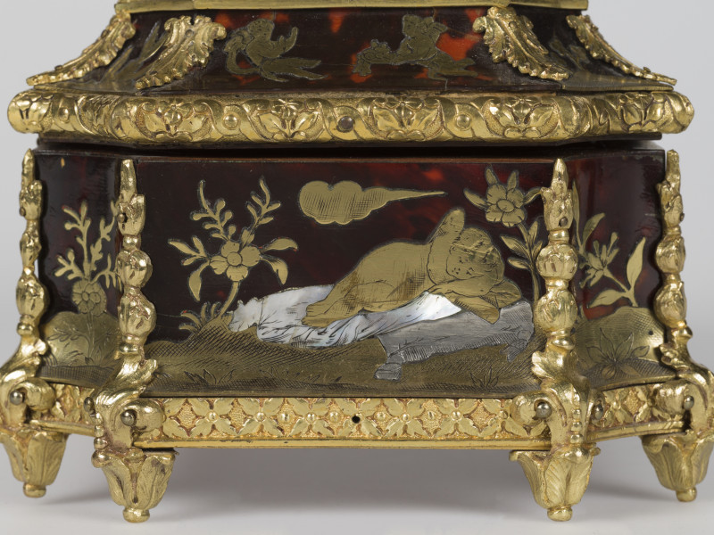 zbliżenie na dekorację z prawego boku - Amor pogrążony we śnie, po bokach motywy roślinne, krawędzie szkatułki pokryte dekoracjami z brązu w postaci palmet i ornamentu kwiatowego, nóżki szkatułki w postaci rozety liści.