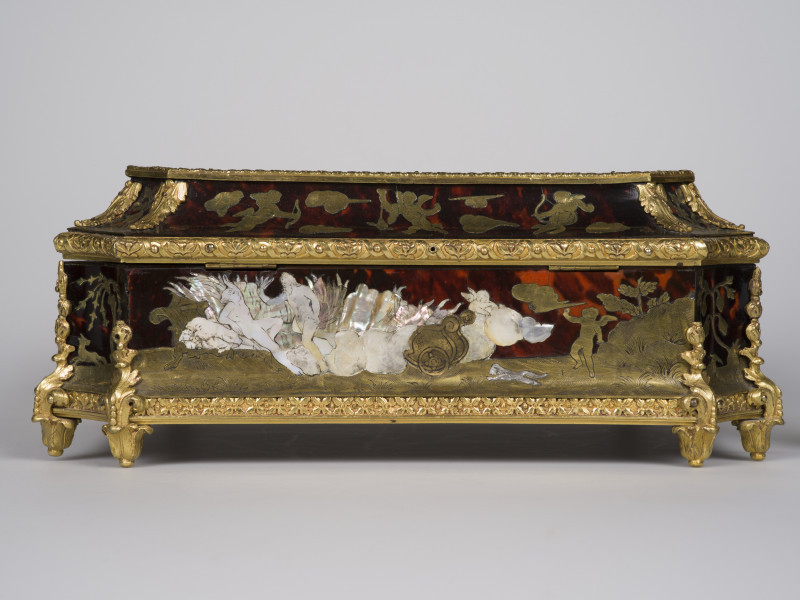 zbliżenie na inkrustowaną dekorację tylnej ścianki, widok z boku. Po środku rydwan i amorek, po prawej złoty amorek na tle wzgórza, po lewej kobieta leżąca na łożu i stojący nad nią mężczyzna, krawędzie ścianek i wieka pokryte dekoracjami z brązu w postaci palmet i ornamentu kwiatowego, nóżki w postaci rozety liści. 